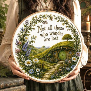 May include: A circular wooden embroidery hoop containing a cross-stitch design of a green hillside with a round door dwelling. The text "Not all those who wander are lost" is stitched in brown above a landscape of wildflowers and a stone path.