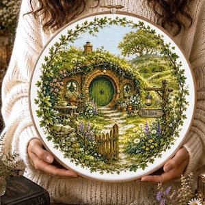 May include: A circular embroidery hoop containing a cross-stitch pattern of a round green door set into a grassy hill. The scene features a stone path, a wooden fence, blooming flowers, and a signpost with the text The Shire written on it.