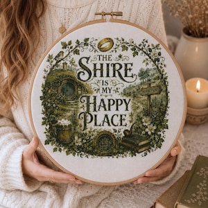 Könnte beinhalten: Ein Kreuzstich-Stickrahmen mit dem Text "The Shire is My Happy Place" und einer detaillierten Illustration eines Hobbit-Loches, einem Schild für Hobbiton und einem goldenen Ring. Das Design ist in Grün-, Braun- und Cremetönen gehalten.