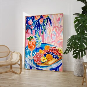 May include: A framed abstract painting depicts a vibrant still life. A table is set with a cocktail, charcuterie, and fruit. The color palette includes blues, pinks, oranges, and greens. The artwork is displayed in a light-colored wooden frame.