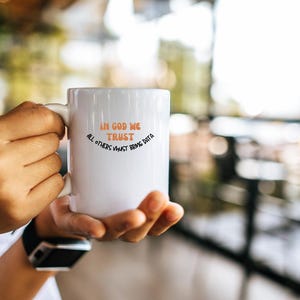 Puede incluir: Taza de cerámica blanca con un diseño en negro y naranja. La taza dice "In God We Trust, All Others Must Bring Data."