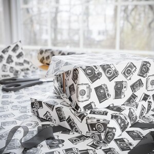 May include: Two gift boxes wrapped in white paper with a black and white vintage camera pattern. The boxes are stacked on top of each other.