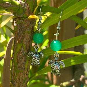 May include: A pair of silver turtle earrings with green jade beads. The earrings are hanging from a plant.