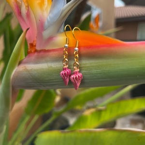 Könnte beinhalten: Ein Paar goldfarbene Ohrringe mit rosa, muschelförmigen Anhängern. Die Ohrringe hängen an goldfarbenen Haken. Im Hintergrund eine farbenfrohe Paradiesvogelblume und grünes Laub.