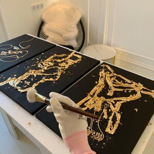 May include: Black canvases with gold leaf horse designs are being created. A person wearing a pink and white glove is using a brush to apply gold leaf. The artwork is on a white table.