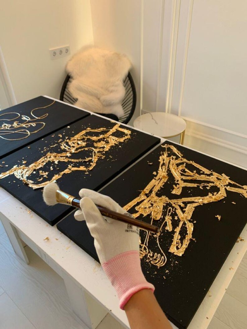 May include: Black canvases with gold leaf horse designs are being created. A person wearing a pink and white glove is using a brush to apply gold leaf. The artwork is on a white table.