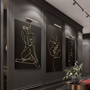 May include: Three abstract line art wall hangings with gold outlines on black backgrounds. The art is displayed in a hallway with dark gray walls, a black marble floor, and accent lighting. The art depicts the female form.