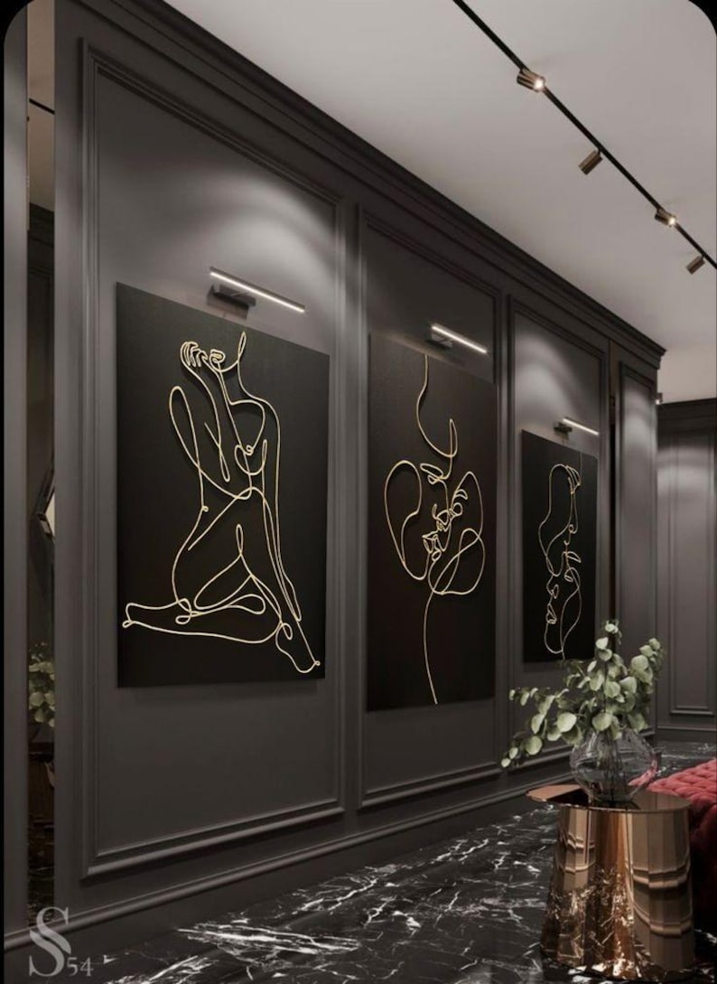 May include: Three abstract line art wall hangings with gold outlines on black backgrounds. The art is displayed in a hallway with dark gray walls, a black marble floor, and accent lighting. The art depicts the female form.