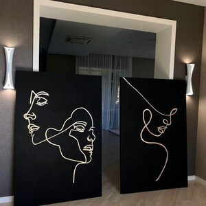 May include: Two black canvas art pieces with gold line drawings of faces. One canvas features two faces, while the other has a single face. The art is displayed in a room with white wall sconces and a doorway.