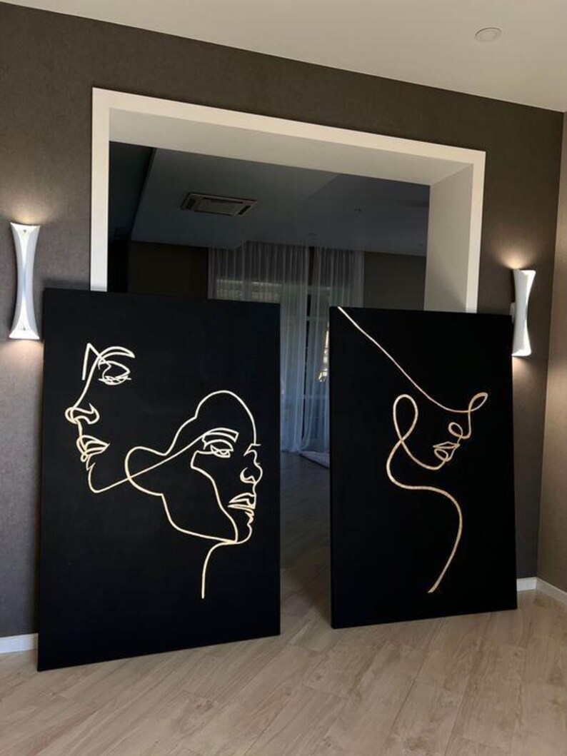 May include: Two black canvas art pieces with gold line drawings of faces. One canvas features two faces, while the other has a single face. The art is displayed in a room with white wall sconces and a doorway.