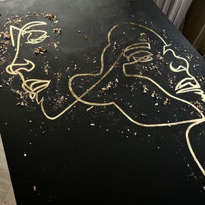 May include: Black artwork featuring gold line art of two faces. Gold leaf flakes are scattered across the surface. The faces are depicted in a minimalist, continuous line style, creating a modern and artistic aesthetic.