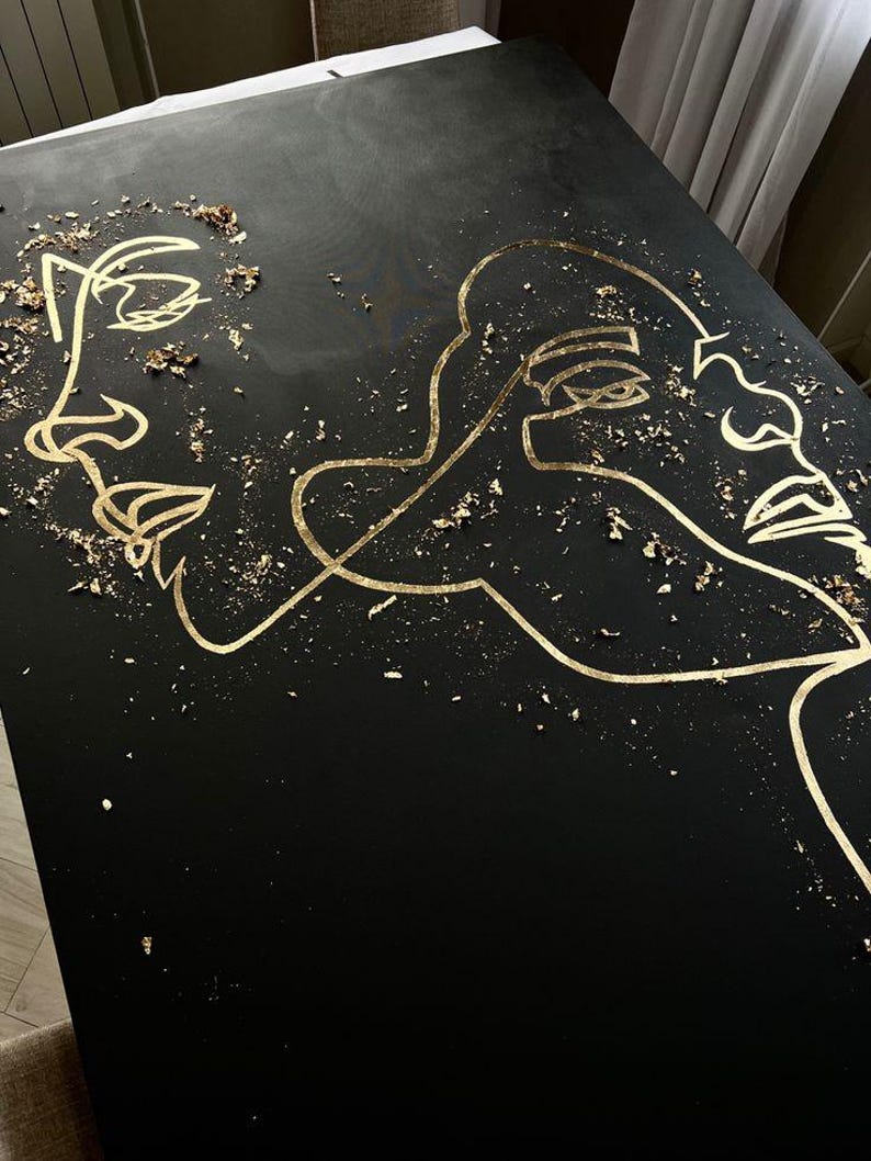 May include: Black artwork featuring gold line art of two faces. Gold leaf flakes are scattered across the surface. The faces are depicted in a minimalist, continuous line style, creating a modern and artistic aesthetic.