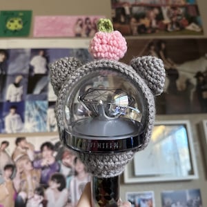 May include: A gray crocheted hat with a pink pom-pom on top. The hat is over a clear plastic sphere with a diamond-shaped light inside. The sphere has the word "SEVENTEEN" printed on it.