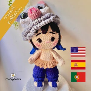 May include: A handmade crochet doll wearing a gray and pink pig-like hat, blue pants, and a tan top. The doll has black hair with blue accents and rosy cheeks. The image includes the text "crochet pattern" and the word "margurumi".