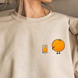 Puede incluir: Una sudadera de color beige claro con un gr&aacute;fico de una naranja exprimiendo jugo de naranja en un vaso. La naranja tiene cara y piernas. El texto "Hoodie & Crewneck in Available Sizes" est&aacute; en la parte inferior de la imagen.