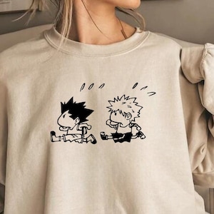 May include: A cream-coloured sweatshirt with a black and white cartoon illustration of two running characters. The text "Hoodie & Crewneck in Available Sizes" is at the bottom. The sweatshirt has a crew neck and long sleeves.