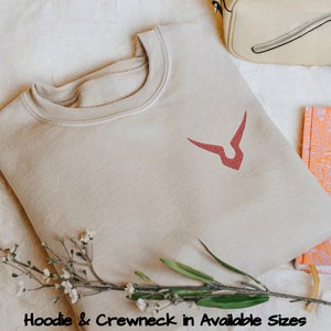 May include: A beige crewneck sweatshirt with a red embroidered symbol on the front. The symbol is a stylised V with a curved line at the bottom. Hoodie & Crewneck in Available Sizes.