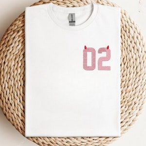 May include: White t-shirt with a pink number "02" with red devil horns on top.