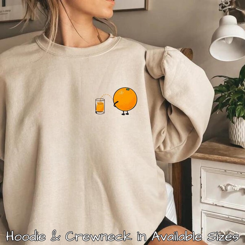 Puede incluir: Una sudadera de color beige claro con un gr&aacute;fico de una naranja exprimiendo jugo de naranja en un vaso. La naranja tiene cara y piernas. El texto "Hoodie & Crewneck in Available Sizes" est&aacute; en la parte inferior de la imagen.