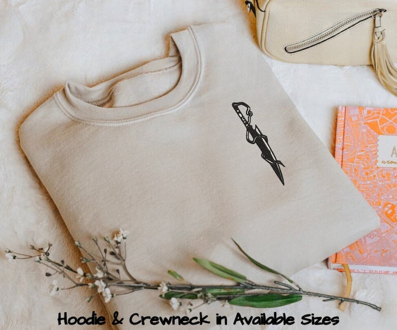 Peut inclure: Un sweat-shirt beige avec un graphique noir d'une &eacute;p&eacute;e avec des vignes enroul&eacute;es autour. Le texte "Hoodie & Crewneck in Available Sizes" est en bas de l'image.