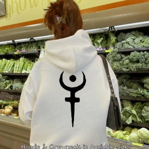 Peut inclure: Un sweat à capuche blanc avec un motif noir au dos. Le motif est un symbole stylisé avec une forme de croissant au-dessus d'un cercle et d'une ligne verticale. Le texte en bas indique "Hoodie & Crewneck in Available Sizes."