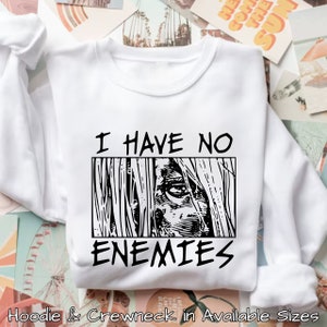 May include: White sweatshirt with black text that reads "I have no enemies" and a black and white illustration of an eye looking out from behind a wall.