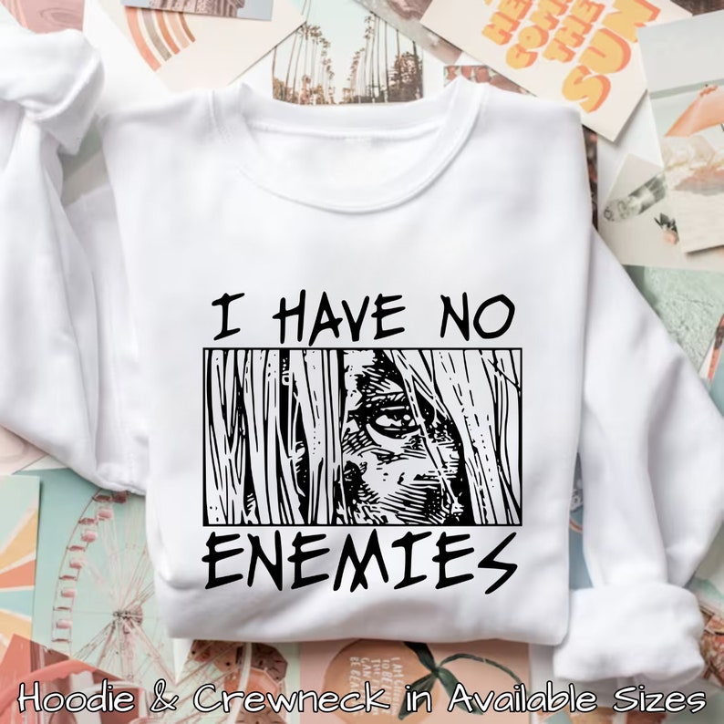 May include: White sweatshirt with black text that reads "I have no enemies" and a black and white illustration of an eye looking out from behind a wall.