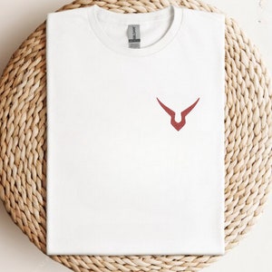 May include: A white t-shirt featuring a small, red emblem on the chest. The emblem is a stylized, angular design. The shirt is made of a soft material and is neatly folded. The shirt is displayed on a woven, light brown surface.