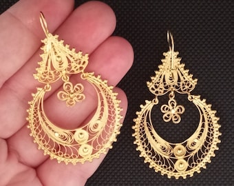 May include: A pair of gold-colored filigree earrings. Each earring features a floral design at the top, a crescent-shaped center, and a small flower pendant. The intricate openwork design is visible.