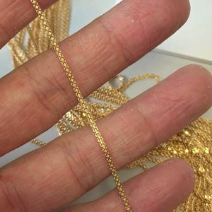 Könnte beinhalten: Nahaufnahme einer goldfarbenen Kette mit filigranem, ineinander verwobenem Design. Die Kette wird zwischen den Fingern gehalten, wodurch ihr kompliziertes Muster und der warme, metallische Farbton zur Geltung kommen. Weitere Ketten sind im Hintergrund zu sehen.