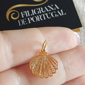 May include: A gold filigree seashell pendant. The pendant is intricately designed with delicate patterns and a small loop for a chain. The background includes a black card with gold text that reads "Filigrana de Portugal."