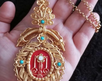 May include: Ornate gold-toned pendant necklace with a red center and turquoise accents. The filigree design features floral and scroll details. The necklace has a delicate chain and is held in a hand.