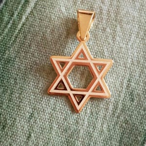 May include: Gold-colored Star of David pendant with a white and brown border. The pendant is suspended from a matching gold bail. The Star of David is a six-pointed star, a symbol of Judaism.