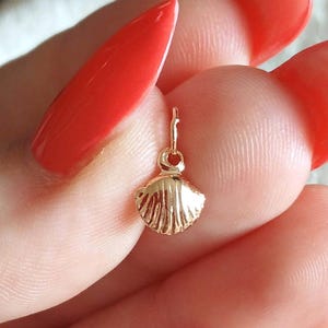 May include: A small, gold-colored seashell charm with a textured surface. The charm has a loop at the top for attaching to a necklace or bracelet. The charm is held in a person's hand.