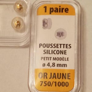 May include: A pair of gold-colored earring backs and a package with the text "1 paire". The package also includes the text "POUSSETTES SILICONE PETIT MODÈLE Ø 4,8 mm OR JAUNE 750/1000".