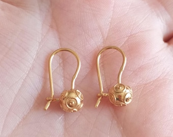 May include: A pair of gold-colored earrings. Each earring features a curved hook and a decorative ball with an intricate pattern. The earrings are displayed against a soft, neutral background.