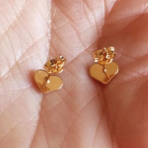 May include: A pair of gold-toned heart-shaped stud earrings with post backs. The earrings feature a smooth, polished surface and are designed with a classic heart silhouette, showcasing a reflective metallic finish on the front and back components.