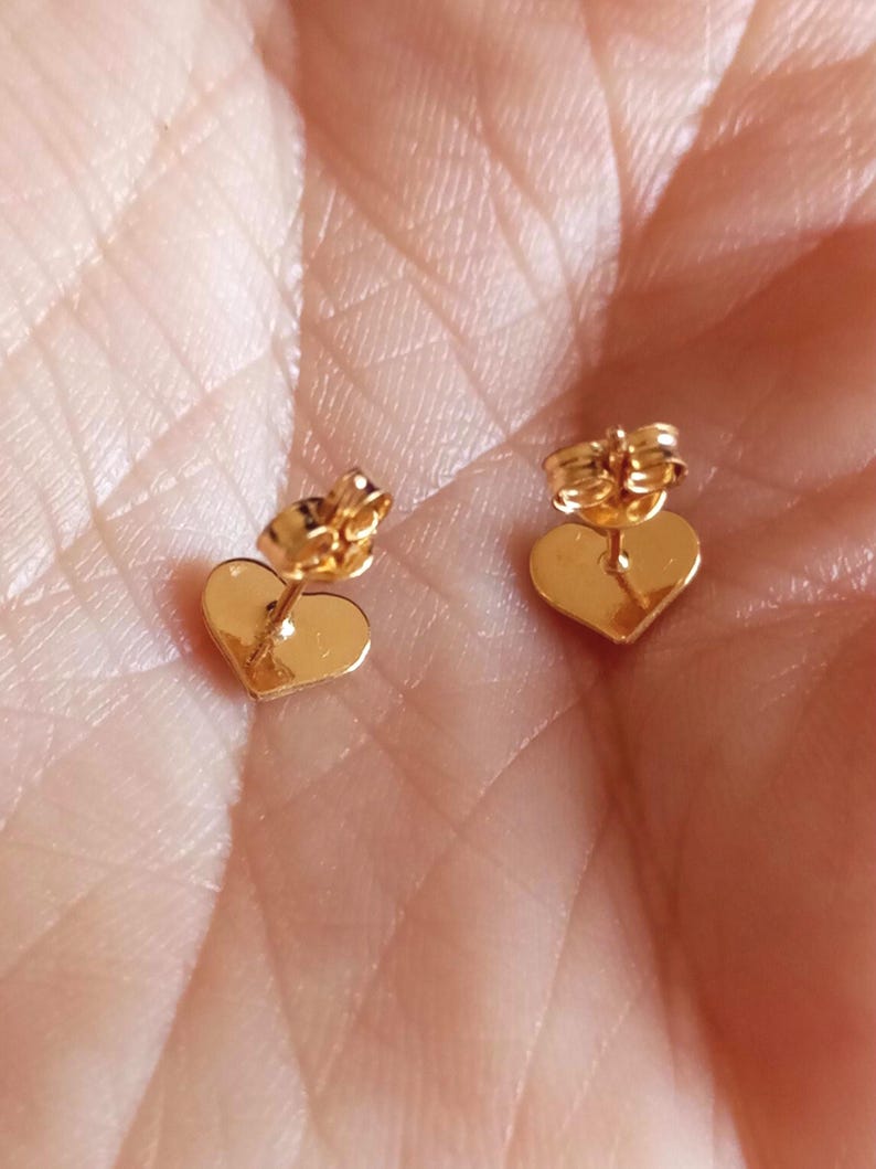 May include: A pair of gold-toned heart-shaped stud earrings with post backs. The earrings feature a smooth, polished surface and are designed with a classic heart silhouette, showcasing a reflective metallic finish on the front and back components.