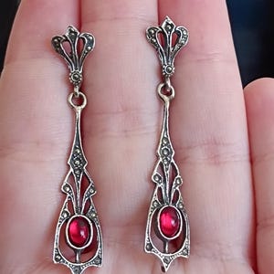 May include: A pair of silver dangle earrings with a red gemstone in the center of each earring. The earrings are decorated with intricate filigree designs and marcasite stones.