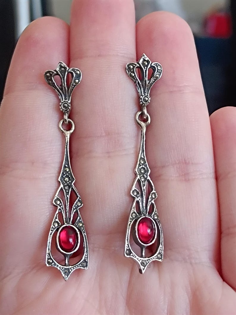 May include: A pair of silver dangle earrings with a red gemstone in the center of each earring. The earrings are decorated with intricate filigree designs and marcasite stones.
