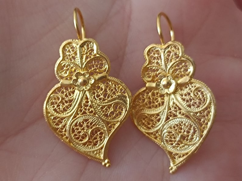 Pu&ograve; includere: Un paio di orecchini a forma di cuore in filigrana d'oro con disegni intricati. Gli orecchini sono delicati e hanno un aspetto vintage.