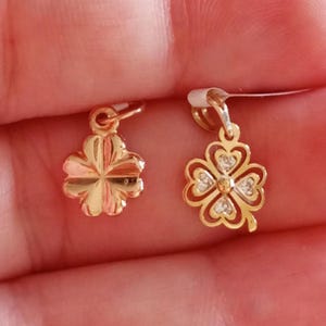 May include: Two gold-colored clover charms. One is a four-leaf clover design, and the other is a four-petal flower. The charms are small and have a loop at the top for hanging. The clover charm has small clear stones.