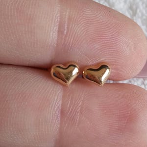 May include: A pair of small, polished gold-tone heart-shaped stud earrings. Each earring features a smooth, rounded surface with a reflective metallic finish, designed as a simple heart silhouette with a pointed base and two rounded lobes at the top.