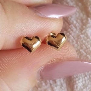 May include: A pair of small, polished gold heart-shaped stud earrings. Each earring features a smooth, rounded surface with a reflective metallic finish, designed as a simple heart silhouette with a post backing for secure attachment to the earlobe.