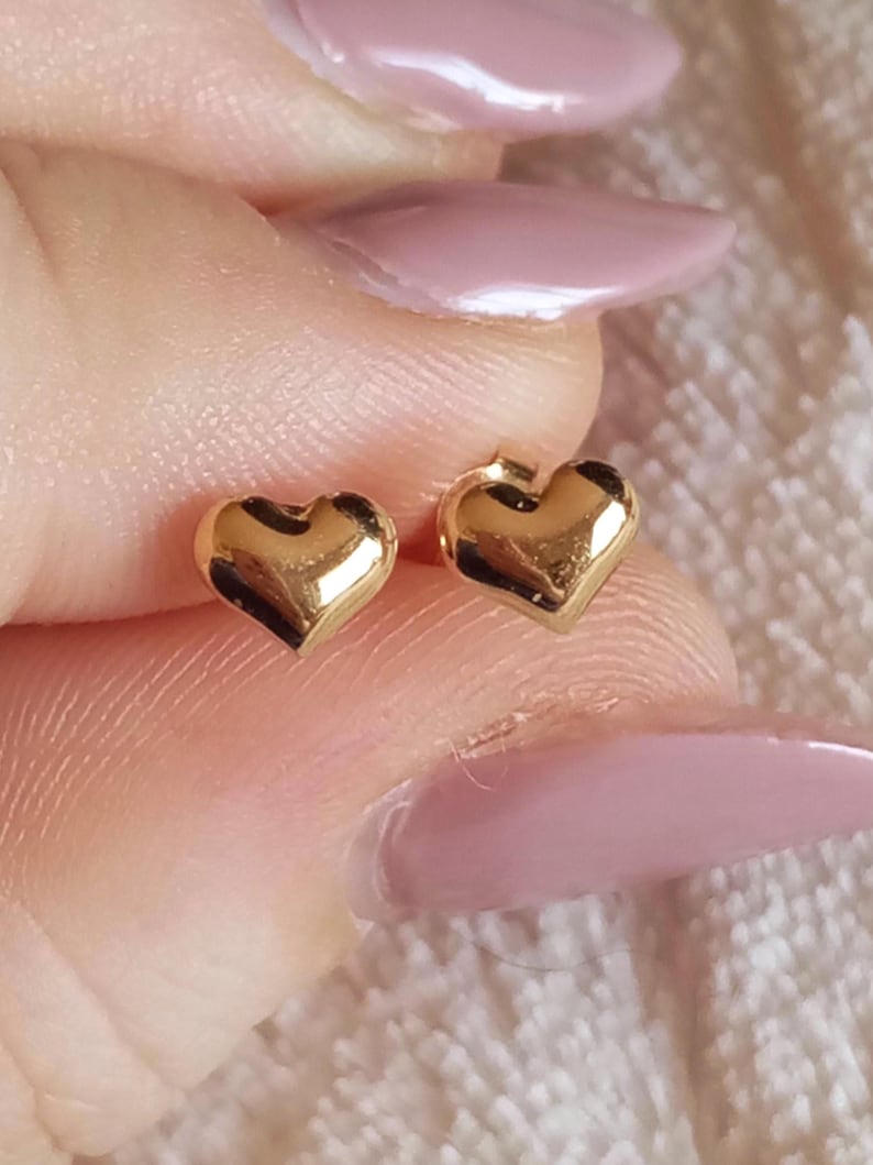 May include: A pair of small, polished gold heart-shaped stud earrings. Each earring features a smooth, rounded surface with a reflective metallic finish, designed as a simple heart silhouette with a post backing for secure attachment to the earlobe.