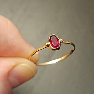 Peut inclure: Une bague en or délicate ornée d'une pierre précieuse ovale rouge vif. La bague a un fin anneau et de petits accents dorés de chaque côté de la pierre. La bague est tenue sur un fond neutre.