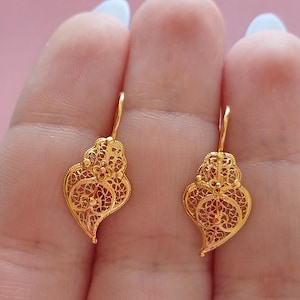 Könnte beinhalten: Ein Paar goldfarbene Ohrringe in Herzform aus filigranem Schmuck. Die Ohrringe haben aufwendige offene Designs und sind an einfachen Haken befestigt. Die Ohrringe werden vor einem weichen rosa Hintergrund gezeigt.