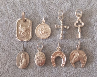May include: A collection of eight silver charms, including a crab, a bull, a cross, a key, a religious figure, a horseshoe, a horse head, and a charm with the text "Agnus Dei".