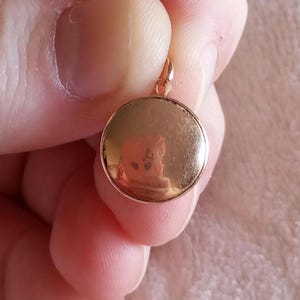 以下が含まれることがあります： 滑らかな表面を持つ丸い金色のペンダント。ペンダントは手で持たれています。