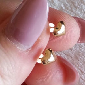May include: A pair of small, polished gold-tone heart-shaped stud earrings. Each earring features a smooth, reflective surface in the shape of a classic heart, designed with a post back for secure attachment to the earlobe.